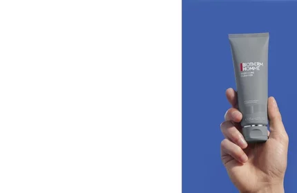 Eine Hand halt eine Biotherm Homme Lait Solaire Tube vor blauem Hintergrund.