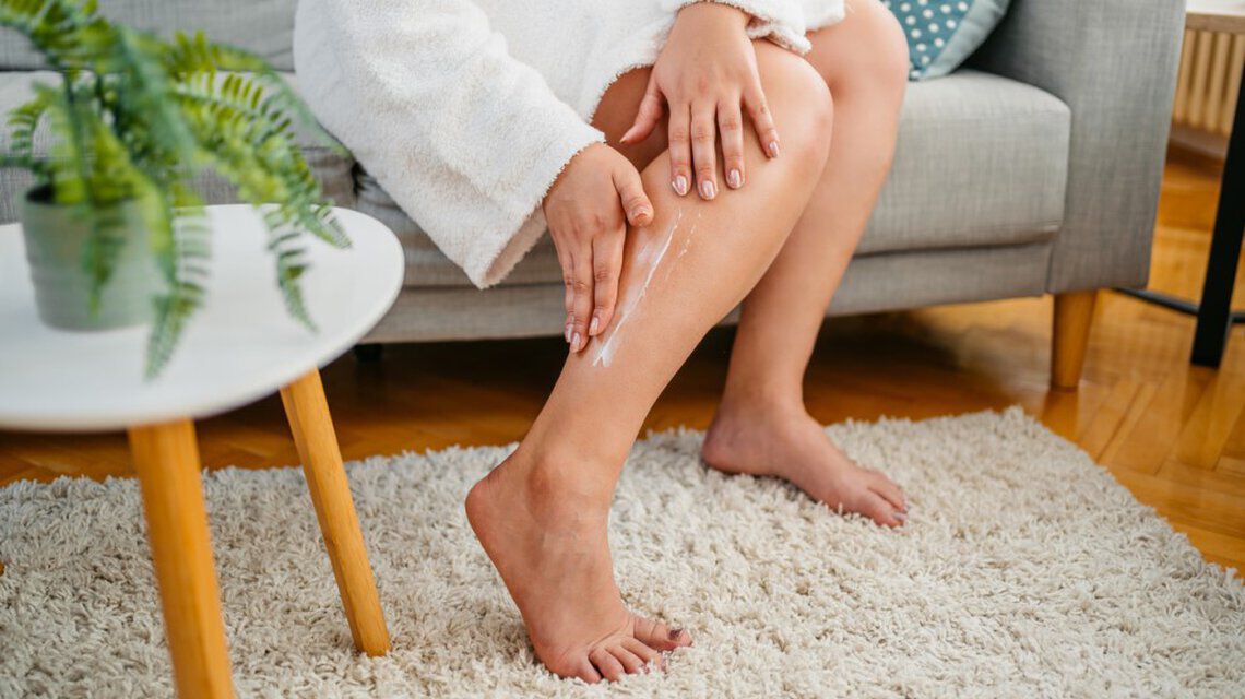 Person cremt Bein ein, sitzt auf einem Sofa im Wohnzimmer, neben einem Tisch mit Pflanze.