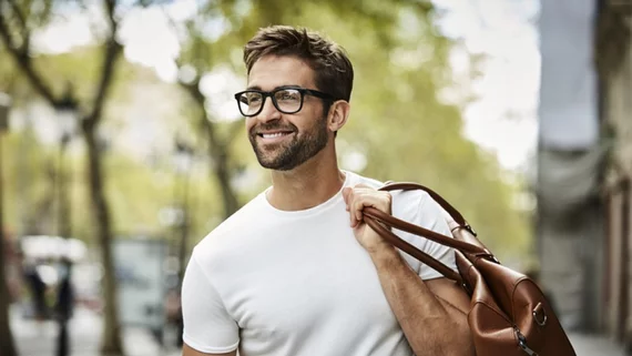Mann mit Brille tragt braune Tasche auf der Schulter; lachelt in einer baumgesaumten, stadtischen Straße.