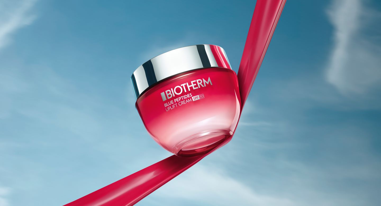 Cremeglas von Biotherm schwebt vor blauem Himmel, gehalten von rotem Band. Text: "BIOTHERM BLUE PEPTIDES UPLIFT CREAM."