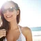 Frau lachelt, tragt Sonnenbrille, steht am sonnigen Strand mit Meer im Hintergrund.