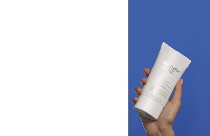 Eine Hand halt eine weiße Creme-Tube vor blauem Hintergrund. Text: BIOTHERM Lait De Gommage.