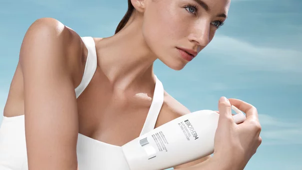 Person halt weiße Lotionflasche mit Text "BIOTHERM Lait Corporel", blauer Himmel im Hintergrund.
