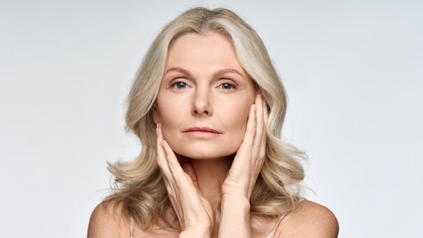 Frau mit blonden Haaren halt ihr Gesicht mit den Handen, neutraler Gesichtsausdruck, weißer Hintergrund.