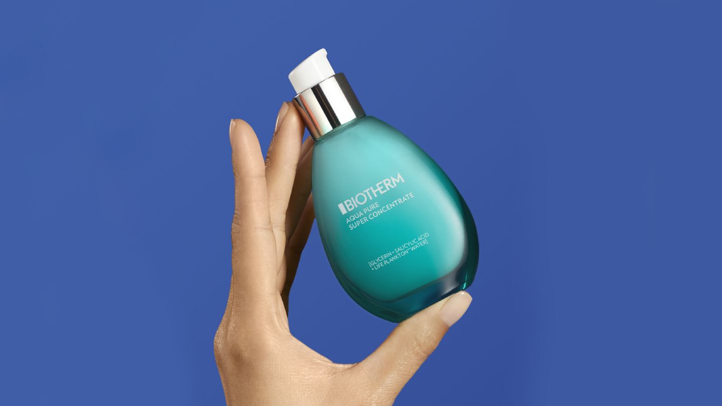 Hand halt eine turkisfarbene Biotherm-Flasche mit Spender vor blauem Hintergrund. Text: "BIOTHERM... REJETON IQ...".