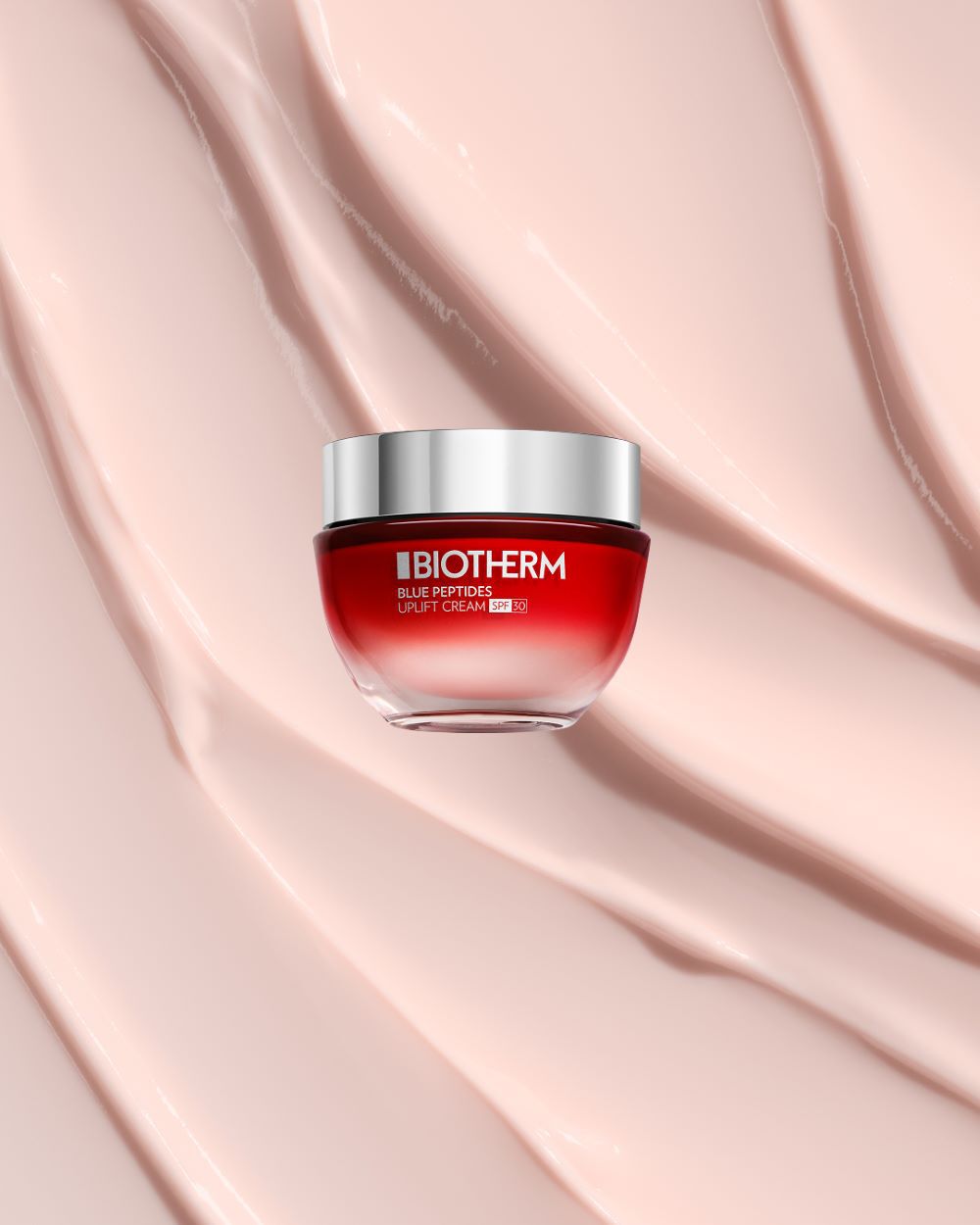 Eine rote Creme-Dose mit silbernem Deckel steht auf einer cremefarbenen, gewellten Oberflache. Text: "BIOTHERM BLUE PEPTIDES UPLIFT CREAM SPF 30".
