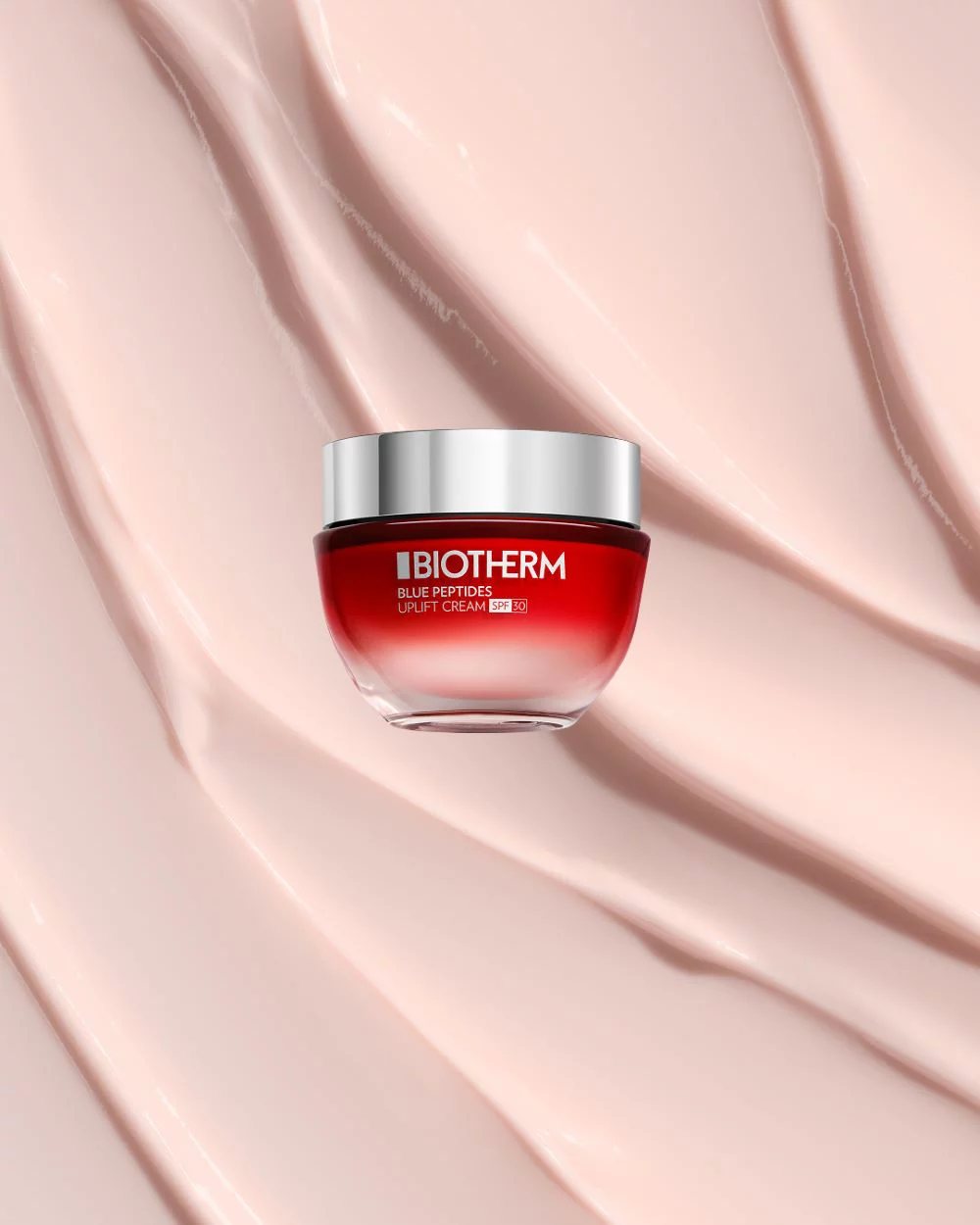 Eine rote Creme-Dose mit silbernem Deckel steht auf einer cremefarbenen, gewellten Oberflache. Text: "BIOTHERM BLUE PEPTIDES UPLIFT CREAM SPF 30".