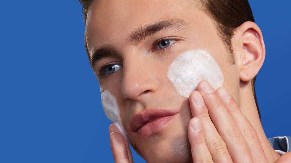 Person tragt Schaum mit den Fingern auf beide Wangen auf; blauer Hintergrund.