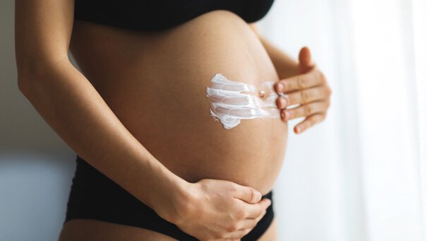 Eine Person tragt Creme auf einen schwangeren Bauch auf, vor einem hellen, unscharfen Hintergrund.