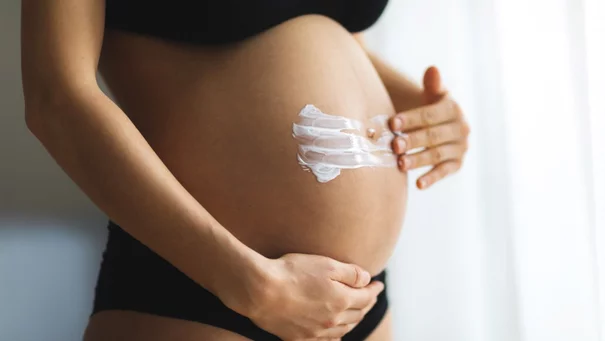 Eine Person tragt Creme auf einen schwangeren Bauch auf, vor einem hellen, unscharfen Hintergrund.