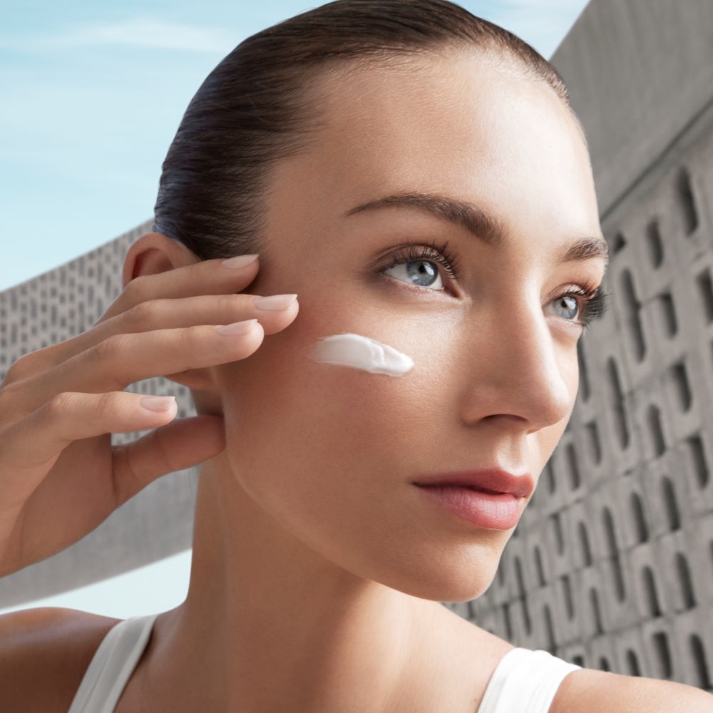 Eine Person tragt Creme auf ihre Wange auf, mit einem modernen Gebaude im Hintergrund.