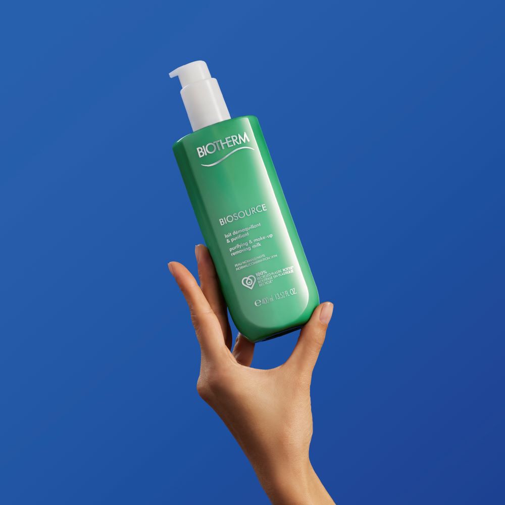 Eine Hand hebt eine grune Flasche mit Pumpverschluss vor blauem Hintergrund.