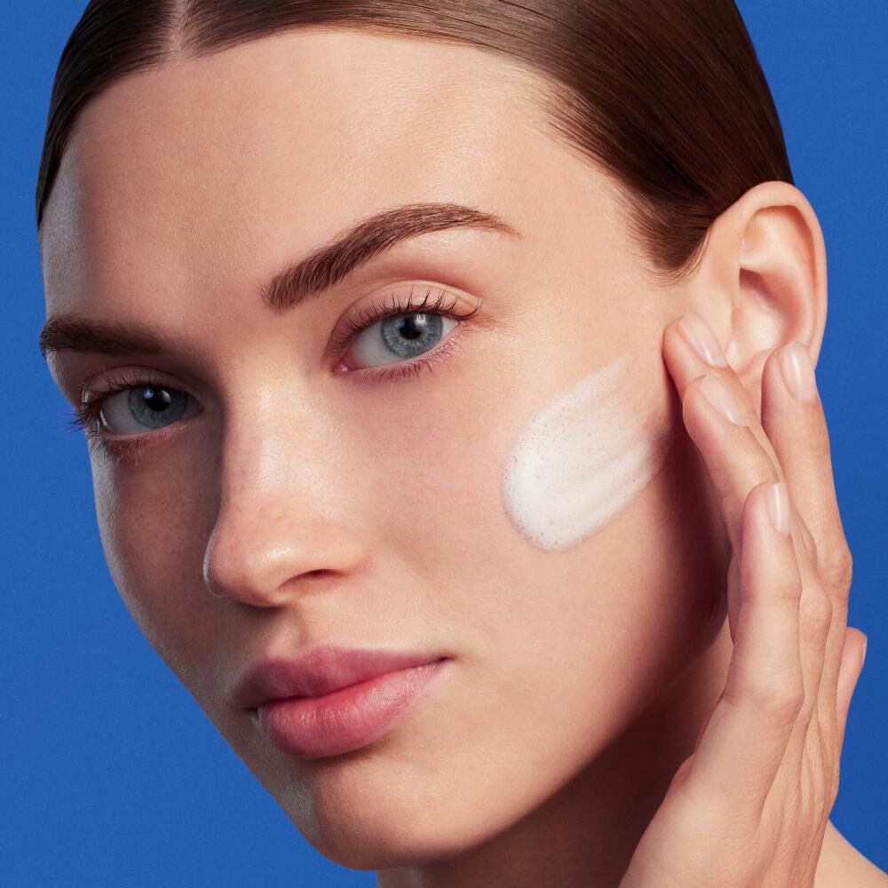 Person tragt Creme auf Gesicht auf, blauer Hintergrund.