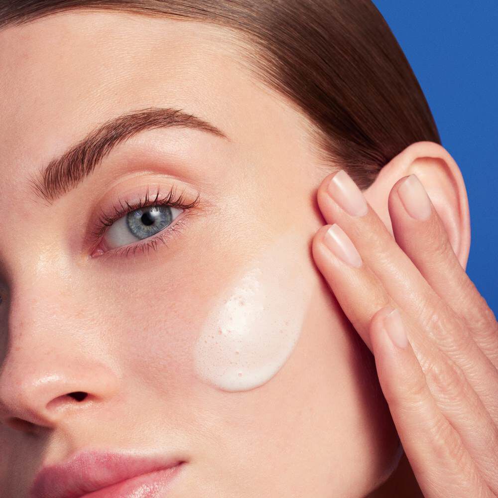 Hand tragt Creme auf Gesicht auf, blauer Hintergrund.