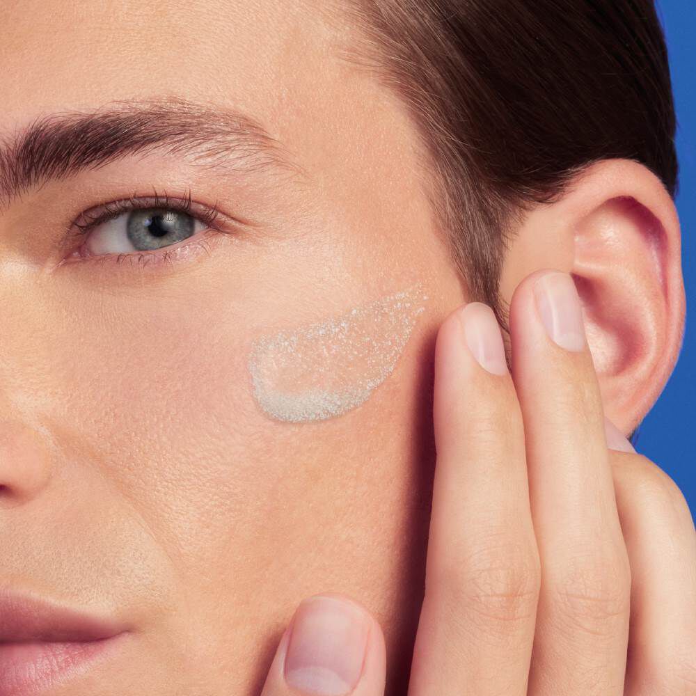 Gesicht mit Creme auf der Wange, Hand beruhrt sanft die Haut vor blauem Hintergrund.