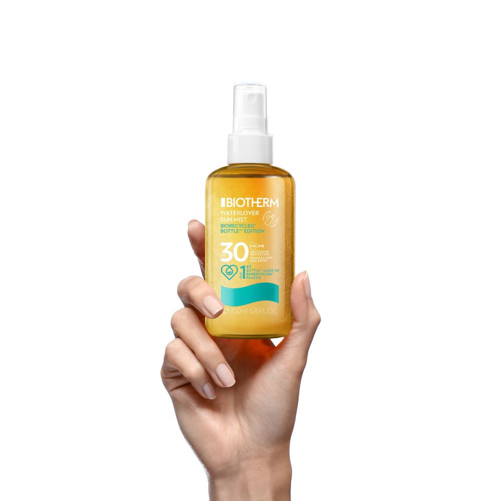 Eine Hand halt eine gelbe Spruhflasche mit orangefarbenem Etikett, vor weißem Hintergrund.