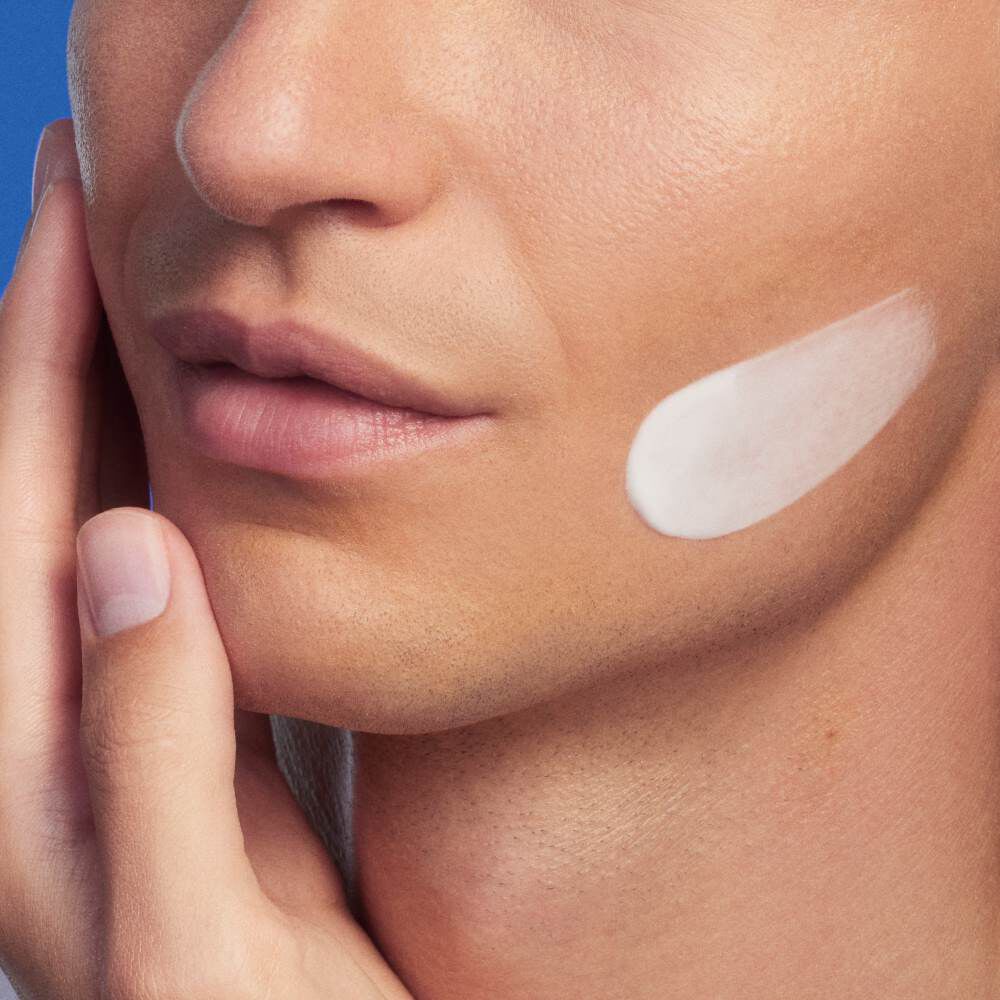 Gesicht mit Creme auf der Wange, Hand beruhrt leicht die Haut, Hintergrund in Blau.