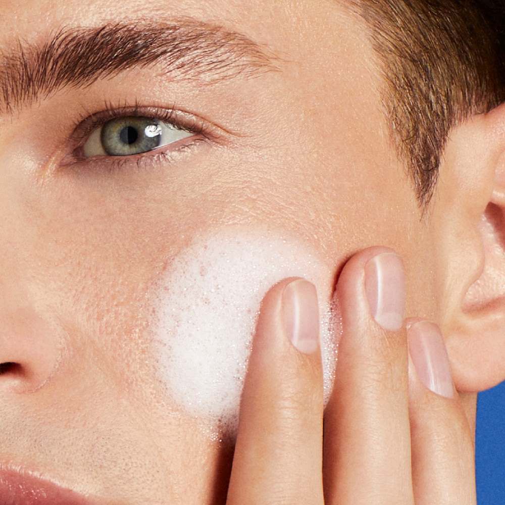 Eine Hand tragt Creme auf eine Wange auf, vor einem blauen Hintergrund.