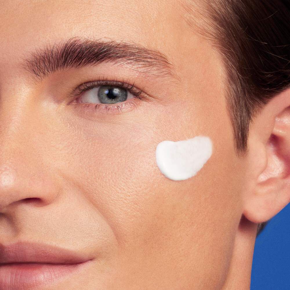 Gesicht mit weißer Creme auf der Wange vor blauem Hintergrund.