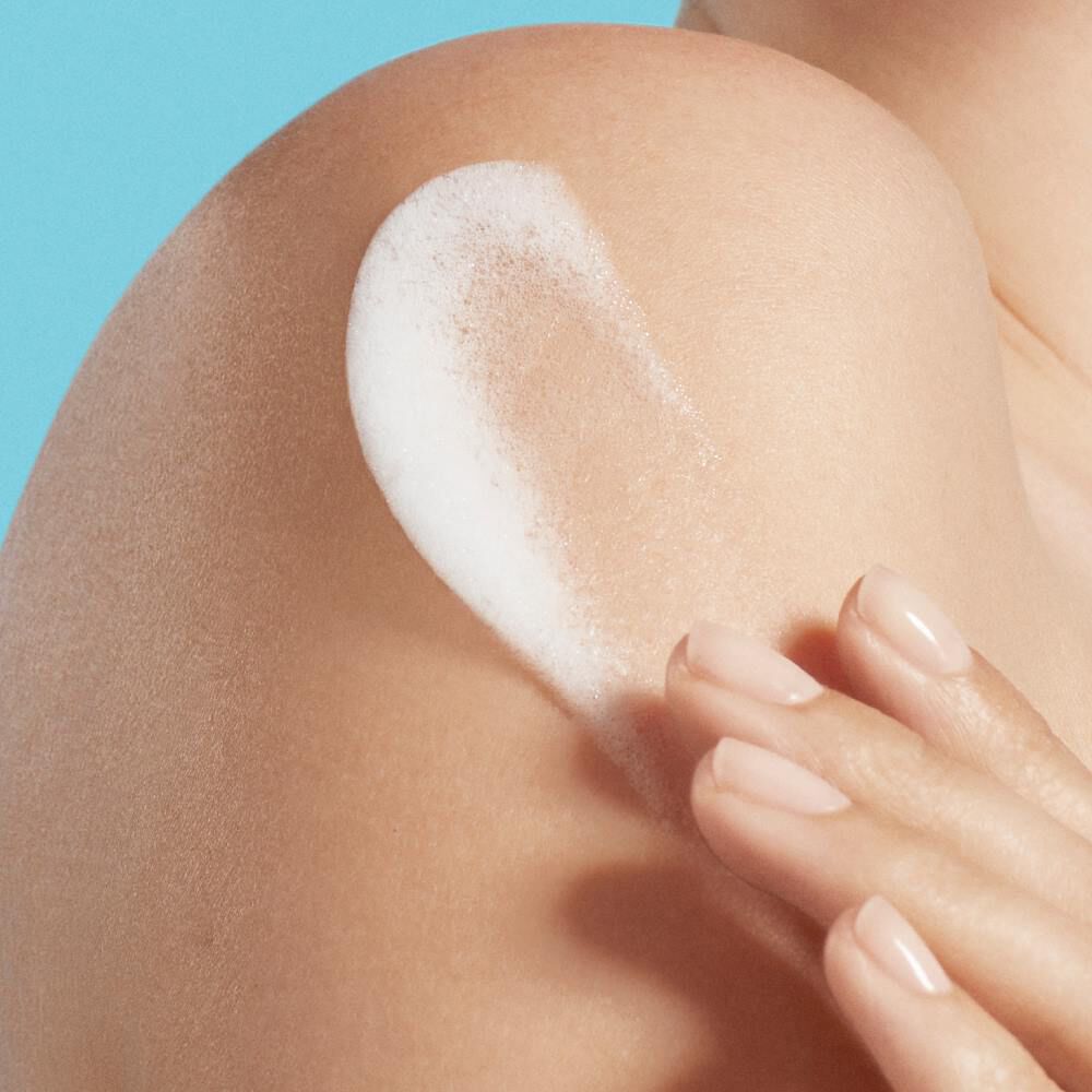 Eine Hand tragt weiße Creme auf eine nackte Schulter auf; blauer Hintergrund.