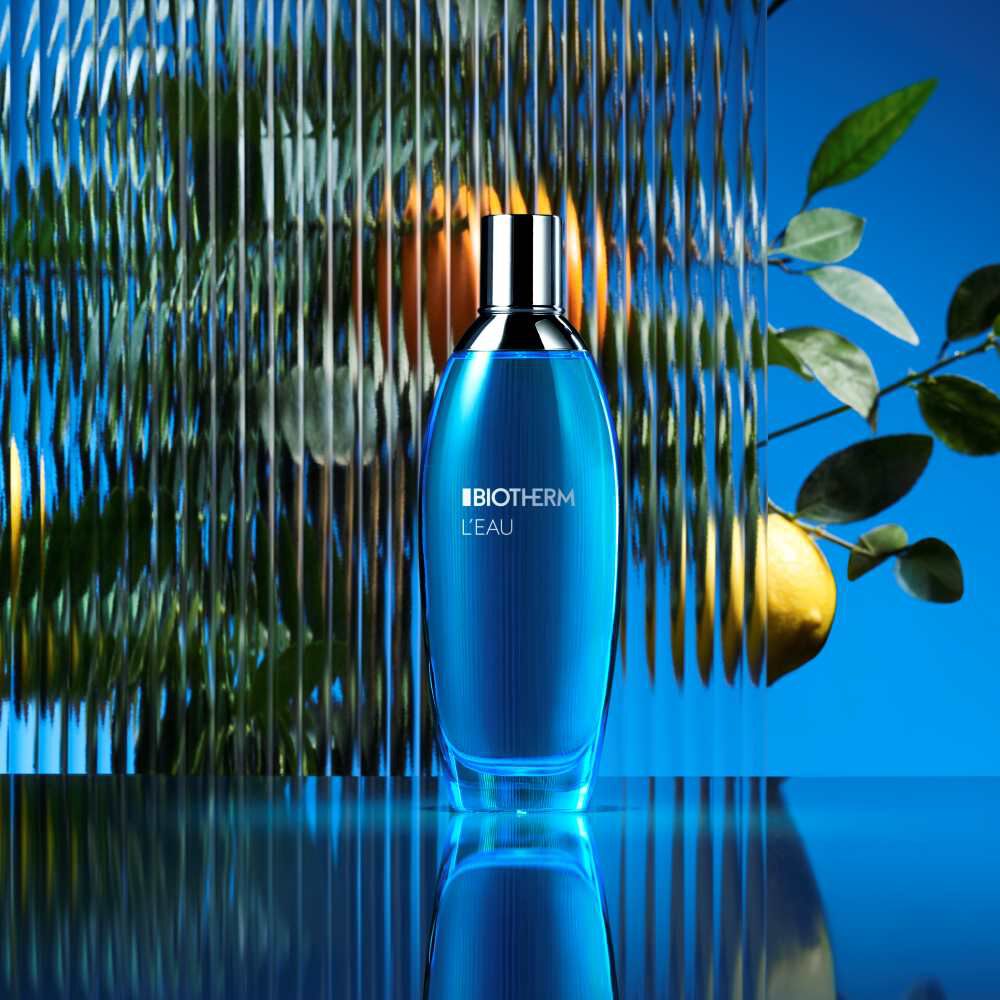 Eine blaue Parfumflasche steht auf reflektierender Oberflache; im Hintergrund Zitrusfruchte.