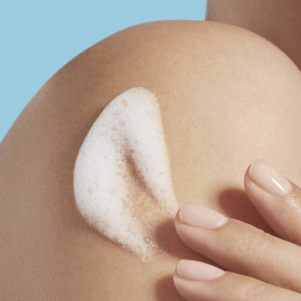 Hand tragt Creme auf Schulter auf, vor blauem Hintergrund.