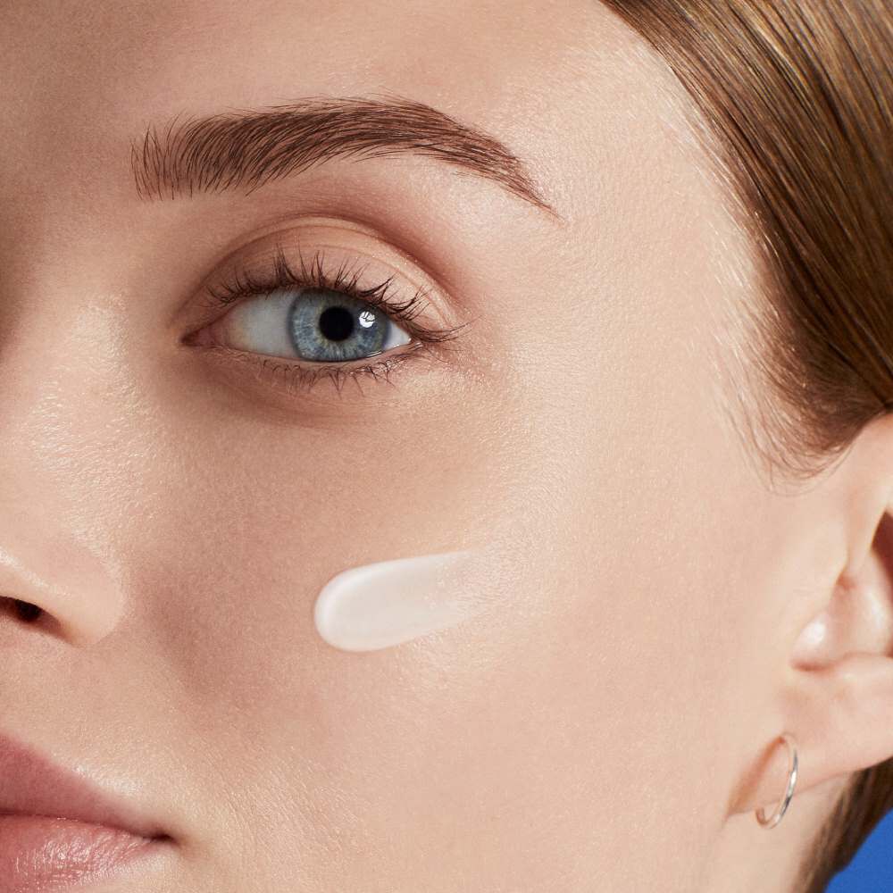 Eine Frau tragt eine weiße Creme auf ihrer Haut auf, Fokus auf Gesichtshalfte, neutrale Umgebung.