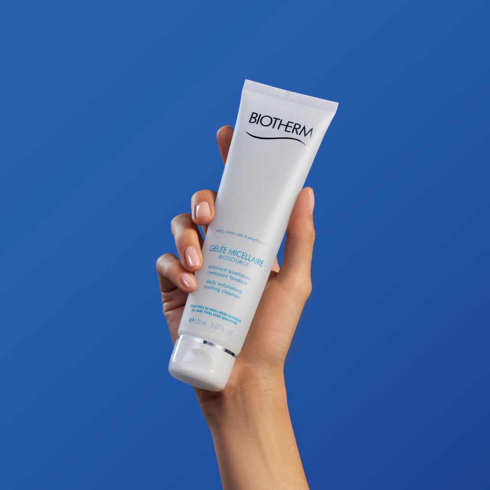 Hand halt eine Hautcremetube vor blauem Hintergrund. Text auf Tube: "CeraVe SA Smoothing Cleanser".